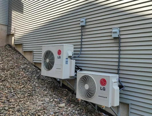 Two ductless mini-split condensers mounted side by side on a gravel bed beside a home
