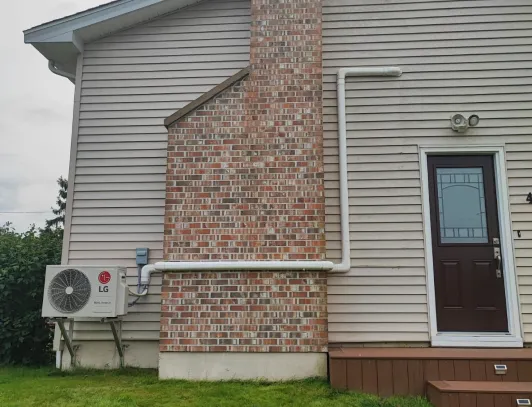 Ductless heat pump condenser installed at the side entrance of a brick and vinyl home