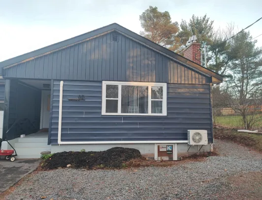 Heat pump condenser installed on a gravel pad beside a dark blue sided bungalow